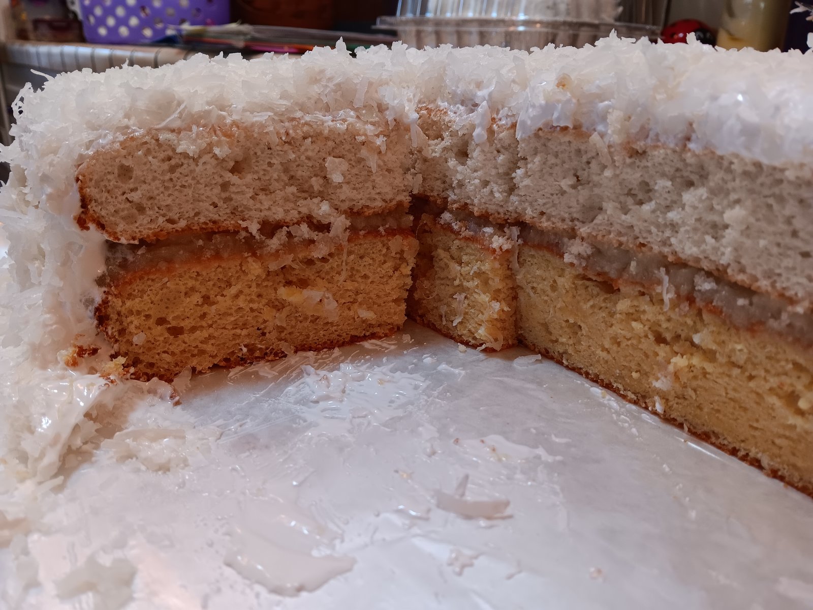 Cut into Royal Hawaiian cake showing white layer, pineapple filling  and yellow layer. All frosted and sprinkled with coconut flakes. 
